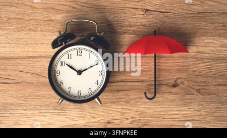 Black vintage alarm clock beside small red umbrella on wooden table time protection and life insurance concept top view. Stock Photo