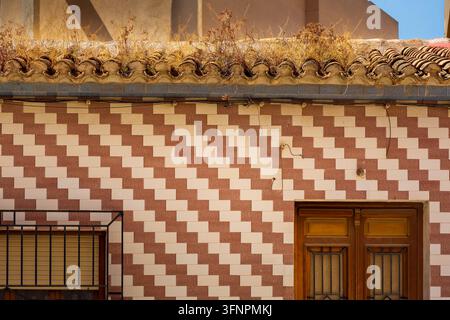 A building wall has red and white square tiles beneath curved brown roof tiles topped with dry weeds. A brown wooden double door with metal bars is on Stock Photo