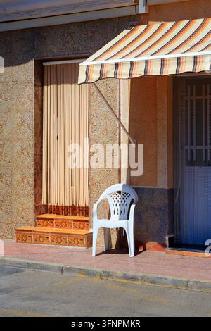 A white plastic chair sits on a red tiled sidewalk outside a building during daylight. Above the doorway hangs a striped awning in orange and brown to Stock Photo