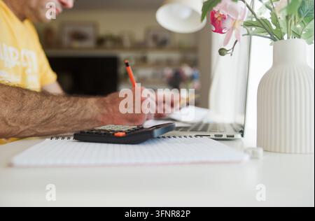 Man calculating finances using a calculator, notebook, and laptop at a home office, managing household budget, paying bills, and reviewing personal ec Stock Photo