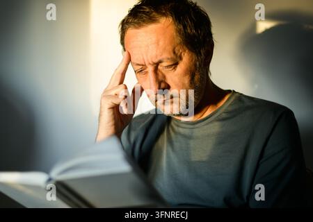 Man absorbed in a book, sitting in a shaft of light with a deep shadow behind him, moody indoor atmosphere. Selective focus. Stock Photo