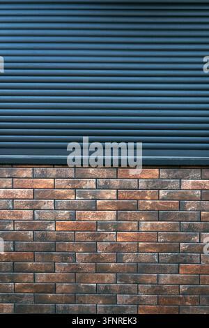 Dark metal roller shutter above a textured brick wall, contrasting horizontal and rectangular patterns, urban architecture detail. Vertical image. Stock Photo