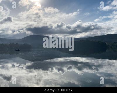 Calm waters reflect a dramatic sky filled with fluffy clouds and a partially hidden sun. A solitary boat is visible on the water, adding a sense of tr Stock Photo
