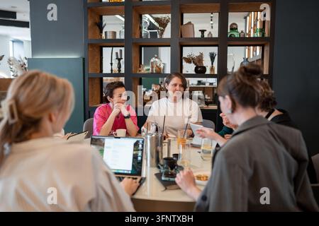 Group of female colleagues enjoying lunch and conversation around round table in modern restaurant, warm lighting, glass partition with frosted Stock Photo
