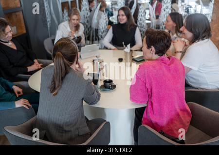 Group of female colleagues enjoying lunch and conversation around round table in modern restaurant, warm lighting, glass partition with frosted Stock Photo