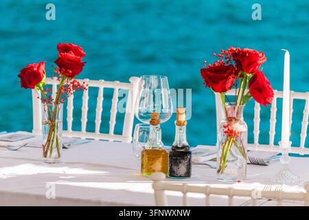 An elegant outdoor table setting with red roses, glassware, bottles, and white chairs overlooks turquoise water. Stock Photo