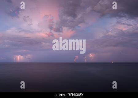 Dramatic lightning strikes across the distant ocean horizon illuminated by a soft purple and pink stormy sunset sky over calm open waters Stock Photo