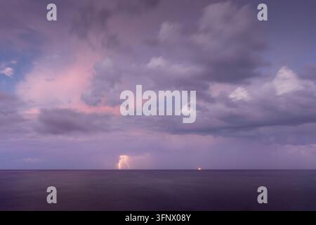 Dramatic lightning strikes across the distant ocean horizon illuminated by a soft purple and pink stormy sunset sky over calm open waters Stock Photo