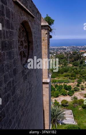 Ancient stone church wall with a carved round window overlooks a green hillside town and distant blue sea under a clear summer sky. Stock Photo