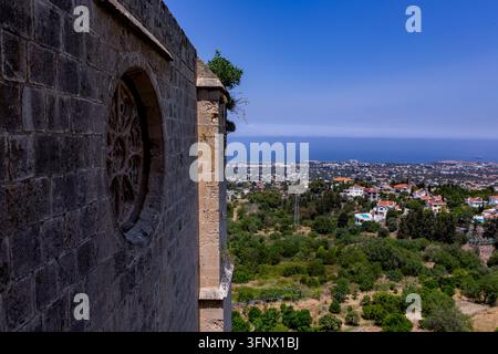 Ancient stone church wall with a carved round window overlooks a green hillside town and distant blue sea under a clear summer sky. Stock Photo