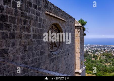 Ancient stone church wall with a carved round window overlooks a green hillside town and distant blue sea under a clear summer sky. Stock Photo