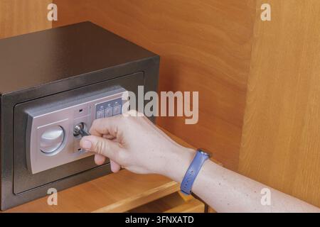 Close up of hand inserting emergency key into keyhole of small black digital safe inside wooden cabinet for unlocking during emergency situation. Stock Photo