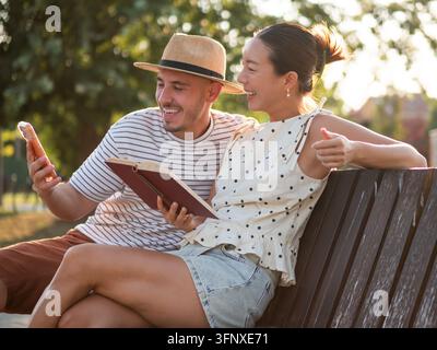 Relaxed woman reading a book while her companion checks his phone during a sunny day in the park. Stock Photo