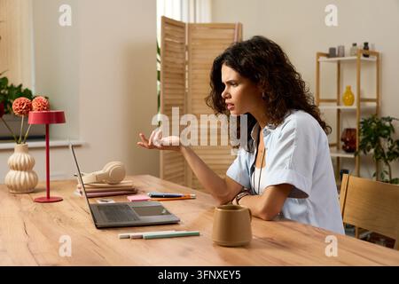 Curly young woman engages with her laptop, having video call in a bright home office space Stock Photo