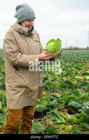 A focused female farmer holds a freshly cut cabbage head, inspecting crop quality in an overcast field. This agricultural worker ensures produce stand Stock Photo