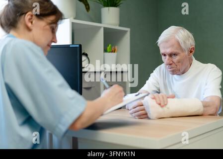 Doctor attentively listening to senior patient during consultation. Elderly man with bandaged hand engaged in conversation with caring professional. Calm environment for clinical communication Stock Photo