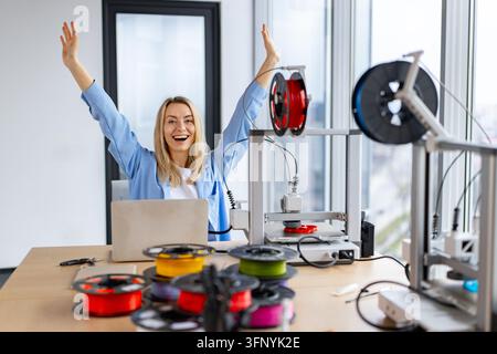 A joyful woman with arms raised in celebration works at a desk with a laptop and multiple 3D printers and filament spools Stock Photo