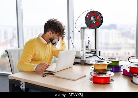 A focused man with curly hair and glasses works on a laptop next to a 3D printer and spools of filament in an office setting Stock Photo