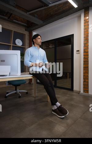 Asian man sitting desk edge holding tablet, wearing light blue shirt and dark trousers in office Stock Photo