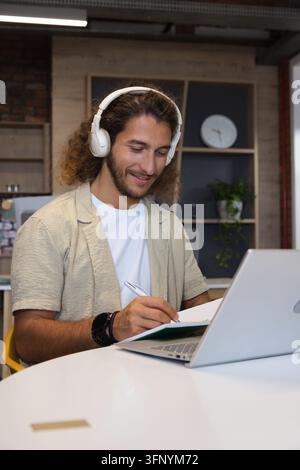 Adult male with white headphones writing in notebook beside silver laptop in study nook Stock Photo