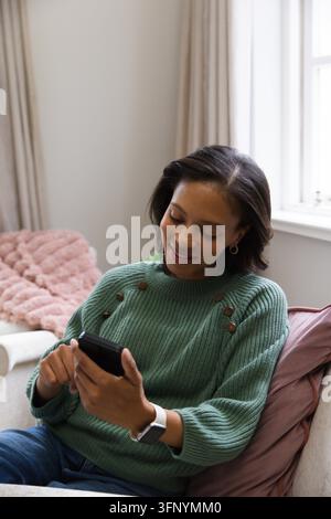 Woman sitting on sofa near sunny window, holding phone and wearing green sweater plus smartwatch Stock Photo