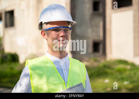 Construction professional wearing hard hat safety glasses and high visibility vest near unfinished building. Architect portrait professional services Stock Photo