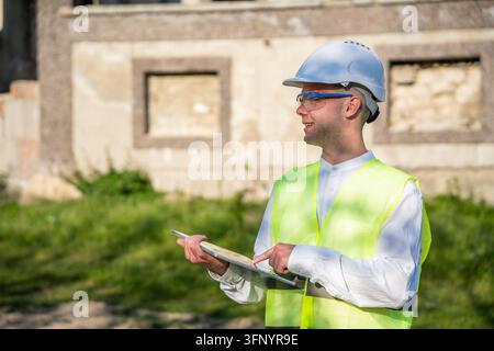 Construction professional using a tablet beside an unfinished building while wearing a hard hat and safety vest. Construction technology digital docum Stock Photo