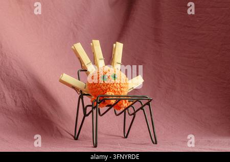 A knitted spider figure sits on a wire base made from clothespins. The sculpture has green eyes and is placed on a pink fabric background. Stock Photo