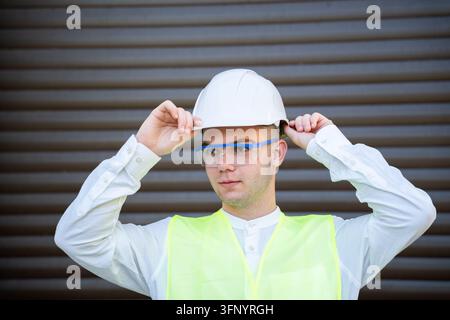 Architect adjusting a white hard hat while wearing safety glasses and high visibility vest before work. Workplace safety personal protective equipment Stock Photo