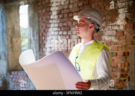 Construction professional in hard hat holding architectural blueprints inside unfinished brick building. Renovation planning design review and buildin Stock Photo