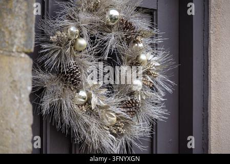 A festive silver glitter wreath adorned with golden baubles pinecones and leaves hangs on a dark brown door. A stone wall is visible on the left. Stock Photo