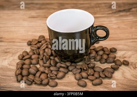 Empty black ceramic coffee cup surrounded by roasted coffee beans on a warm wooden tabletop, with visible grain and natural rustic textur Stock Photo