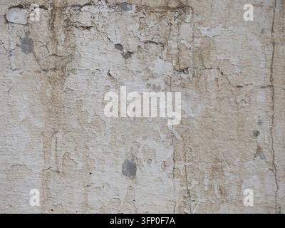 Old grungy cement wall surface featuring cracked beige stucco layers, exposed concrete spots, and dark water drip marks. Stock Photo