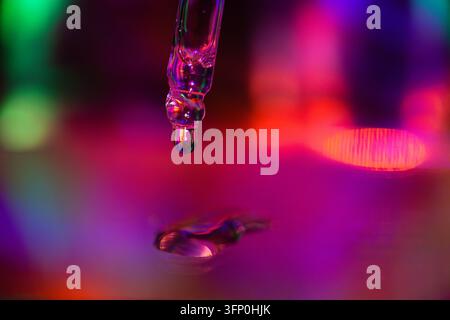 Glass pipette releases a water droplet above a reflective surface, surrounded by blurred magenta, blue, green, and orange light reflections in macro v Stock Photo