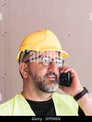 Close-up portrait of a building surveyor in yellow hard hat and reflective vest talking on a smartphone during work on an unfinished construction site Stock Photo