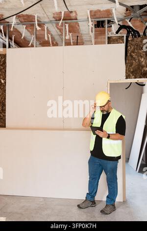 Building surveyor in hard hat and reflective vest checks a digital tablet inside an unfinished room with drywall, exposed ceiling structure and insula Stock Photo