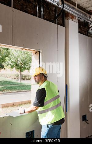 Building surveyor uses a clipboard beside an unfinished window opening while checking site measurements, drywall and exposed building services during Stock Photo