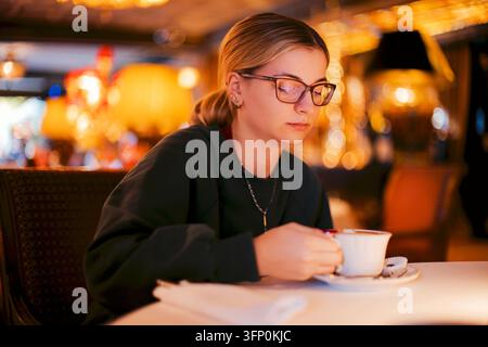 Young woman in glasses sits at a cafe table, holding a white cup while warm blurred lights and softly illuminated furnishings fill the background Stock Photo