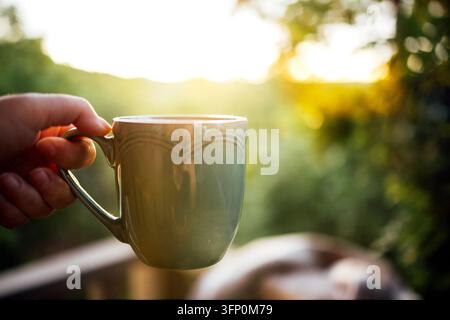 Hand holds a decorative ceramic cup near a sunlit railing, with softly blurred green hills, trees, and golden light in the background Stock Photo