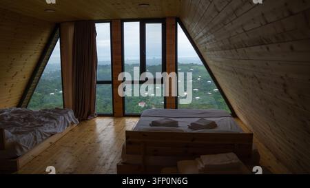 A-frame cabin bedroom interior presenting a fresh bed and stacked towels, framed by large windows revealing a scenic mountainous landscape with scatte Stock Photo