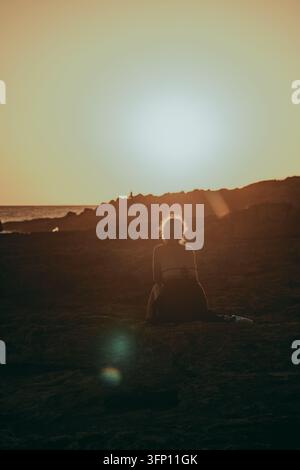 A woman sits on a rocky beach, her back to the viewer, silhouetted against a golden sunset in Mallorca, Spain. The low sun creates a warm, backlit atm Stock Photo