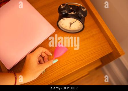 Hand with colorful manicure holds a pink menstrual cup beside stacked pink papers and a vintage alarm clock on a wooden desk surface Stock Photo