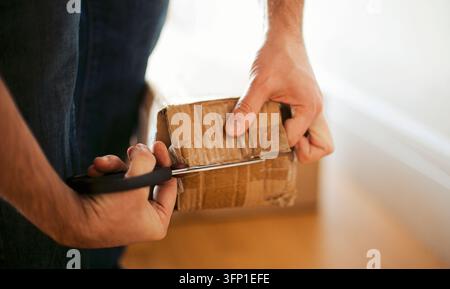 Person uses scissors to cut tape on a small cardboard parcel while holding it beside a bright window above a wooden surface Stock Photo