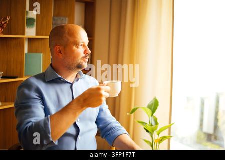 Bald man in blue shirt holds a white cup beside a leafy houseplant, seated near bright window light and wooden shelving in a home interior Stock Photo