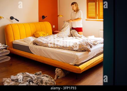 Woman arranges a patterned duvet on a yellow upholstered bed beside a sunlit window, with bedside lamps, wooden flooring, and scattered bedding Stock Photo