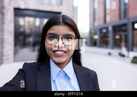 Young indian businesswoman in glasses and business attire standing outdoors in an urban setting, smiling confidently while capturing a selfie, reflecting professionalism and modern connectivity Stock Photo