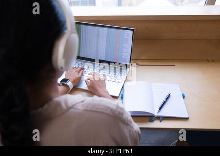 Indian woman wearing headphones, typing spreadsheet on laptop at window counter, copy space Stock Photo