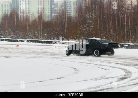 Black car drifting on snowy track, urban practice session with visible tire marks, overcast sky, apartment blocks in background, Stock Photo