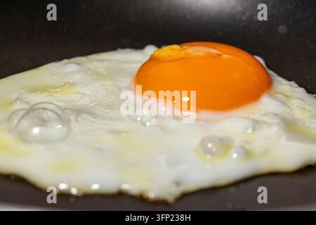 Close-up of a fried egg cooking in a frying pan with glossy orange yolk, bubbling egg white and crispy edges Stock Photo