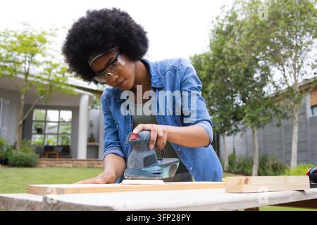 Woman operating orbital sander on wooden plank at yard bench wearing goggles and denim shirt Stock Photo